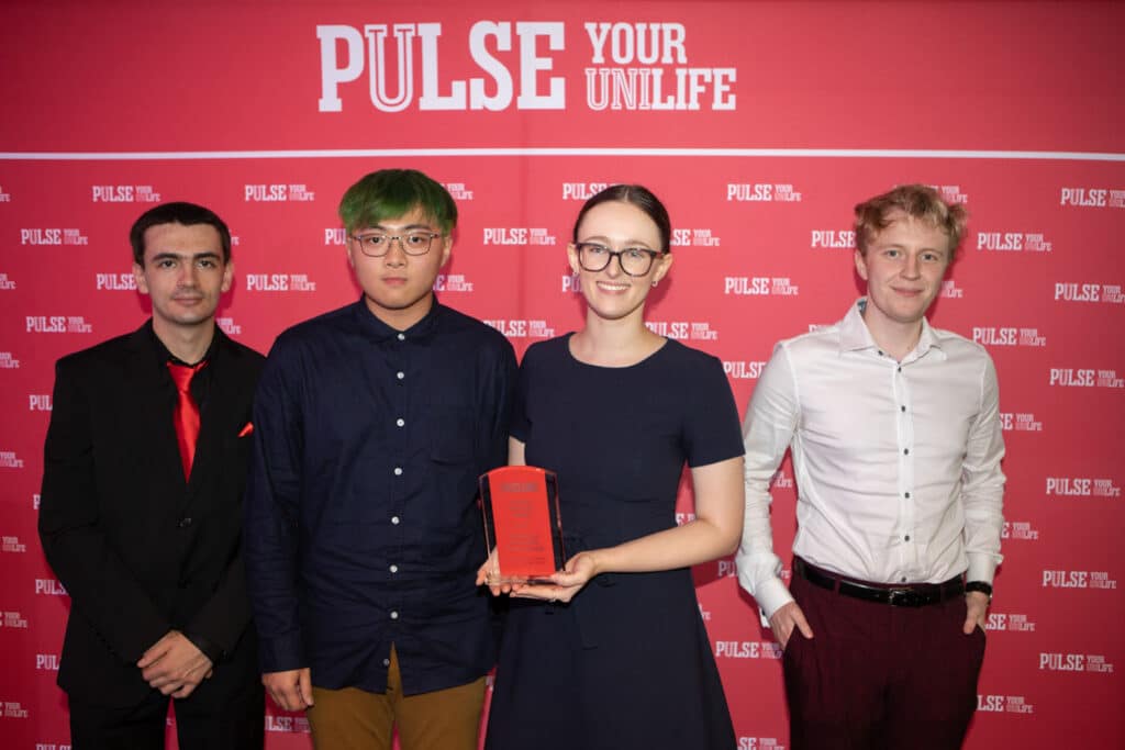 4 people, 1 holding a red award in front of a red and white backdrop
