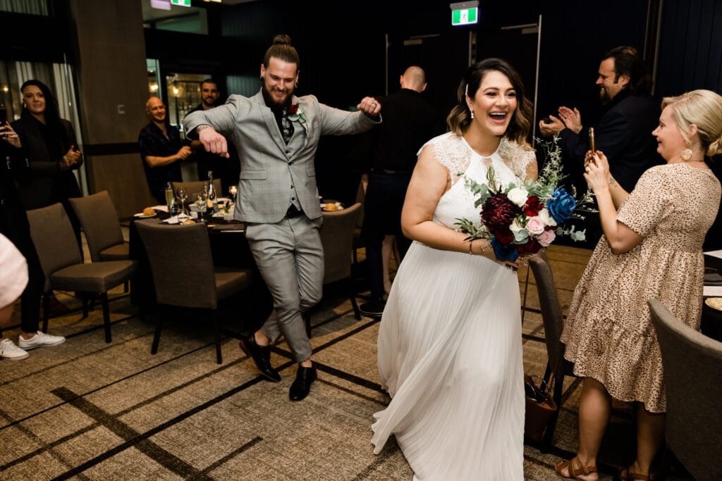 A bride and groom entering a room smiling with an audience standing up