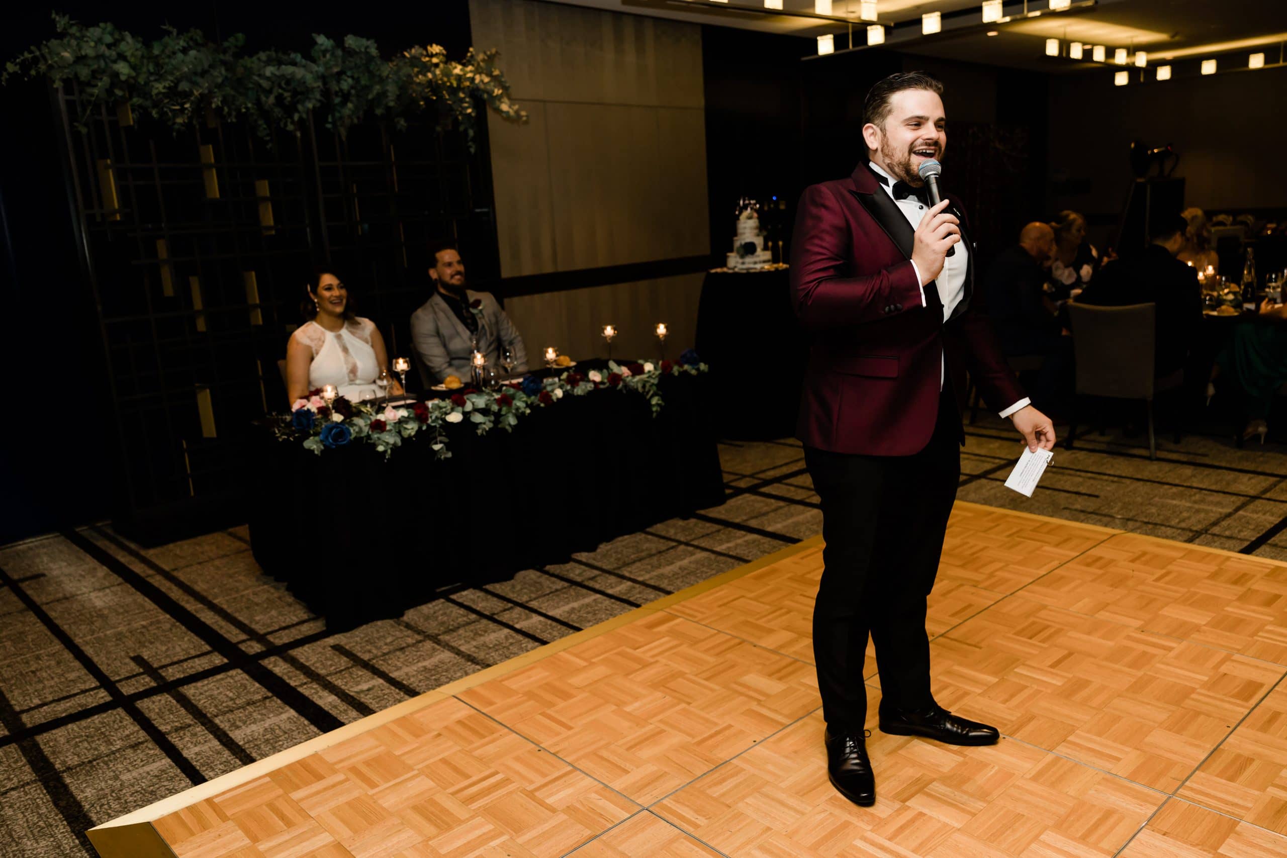 Nathan Cassar smiling and speaking into a microphone with a bride and groom sitting behind