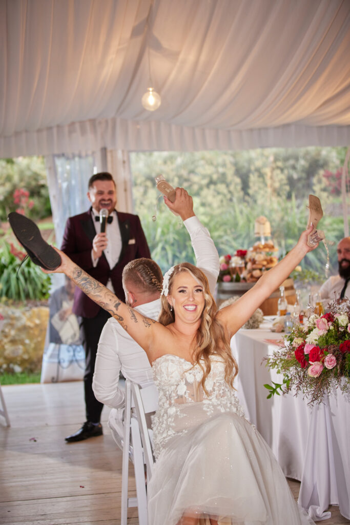 Bride and groom sitting down with their shoes in the air. Nathan Cassar, professional MC is in the background talking on a microphone.
