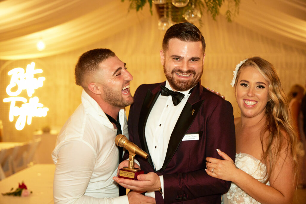 Nathan Cassar, professional wedding mc, standing in the middle of a bride and groom in a red tuxedo and holding a golden microphone trophy. The groom on the left is smiling excitedly and turned to the centre, the bride is smiling and holding Nathan's arm.