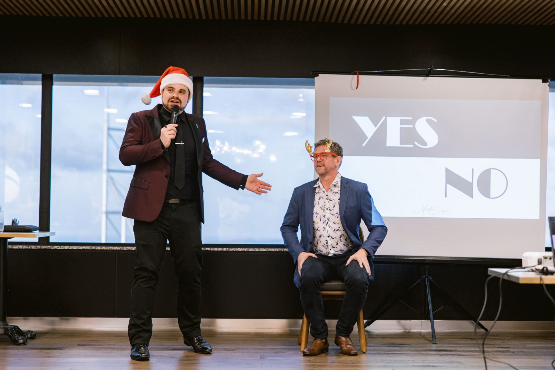 Nathan Cassar, corporate MC standing on stage in a red tuxeo jacket and wearing a santa hat with a guest sitting down on a chari to his right with a screen in the background that reads "YES NO"