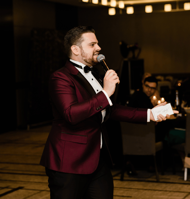 Nathan Cassar, MC in a red tuxedo holding a microphone and palm cards at a wedding