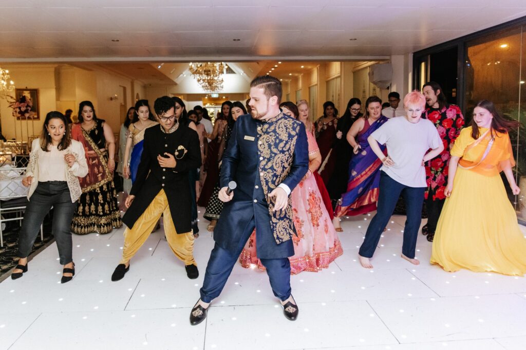 Nathan Cassar, MC dressed in a fancy blue and gold Indian sherwani holding a microphone in front of a crowd of people in lines dancing behind him on a dancefloor at a wedding