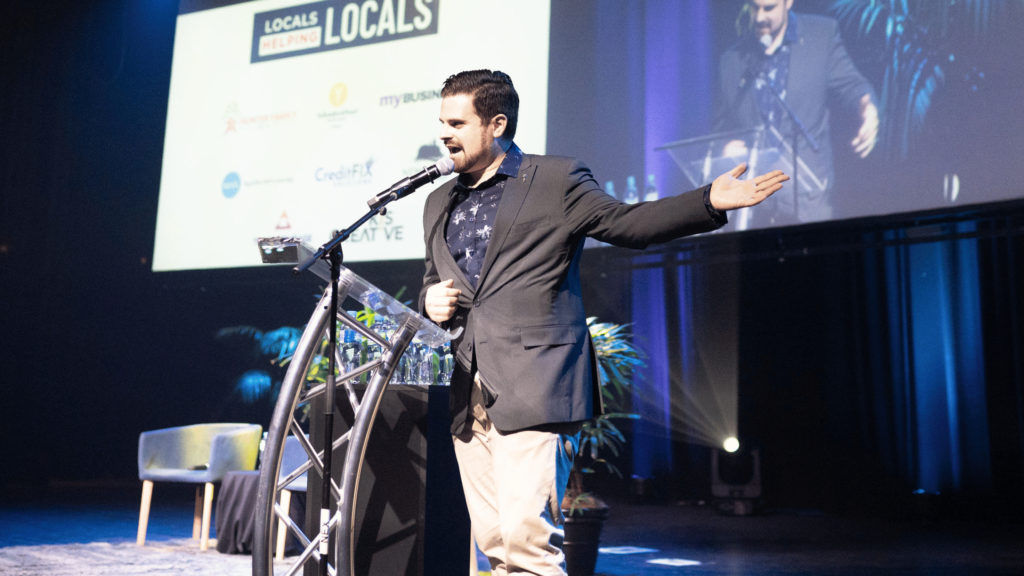 Nathan Cassar MC onstage speaking into a lectern and pointing to the right.