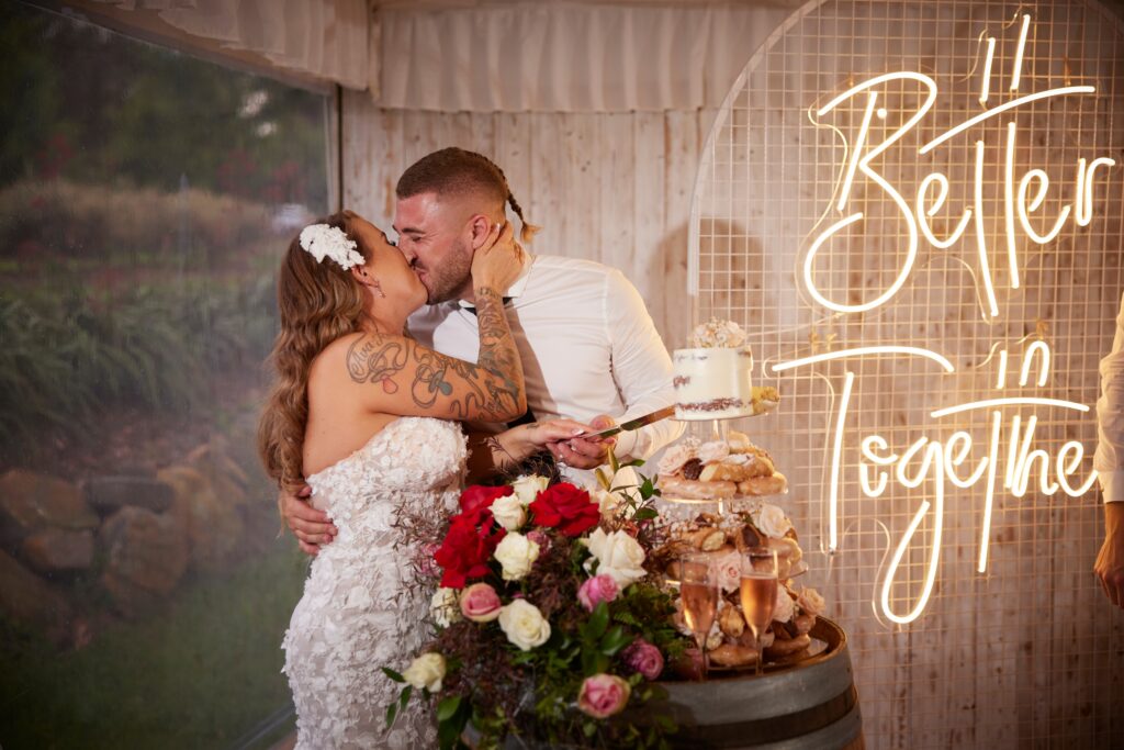 Newlyweds, Allison & Ben, kissing after cutting their cake in front of a neon sign that reads "Better Together"