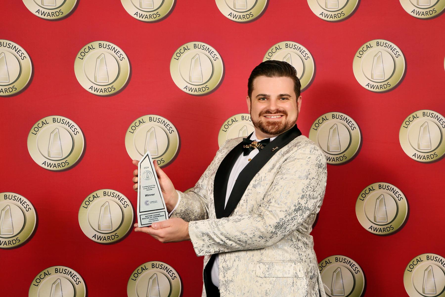 Nathan Cassar Master of Ceremonies, winner of the Cumberland City Local Business Awards 2024 Sole Operator of the Year, holding his award trophy at Merrylands RSL in a white tuxedo made by Woolcott St