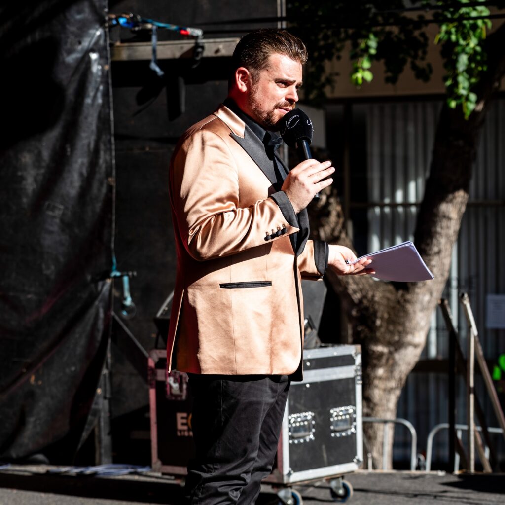 Nathan Cassar, corporate event mc in gold jacket speaking on outdoor stage