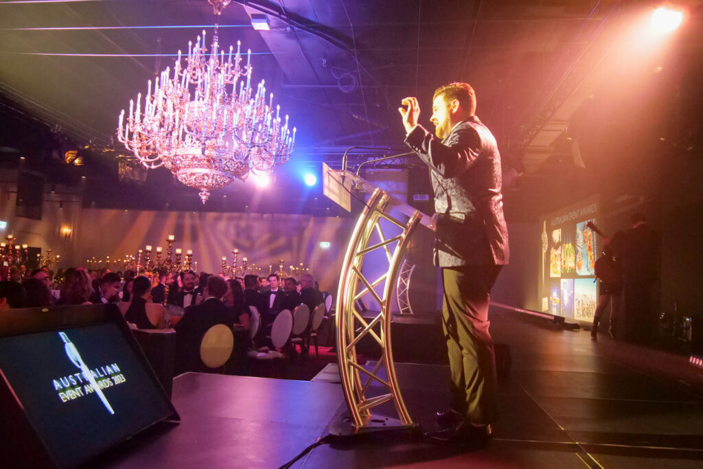Nathan Cassar, corporate event mc speaking at formal awards ceremony under chandelier
