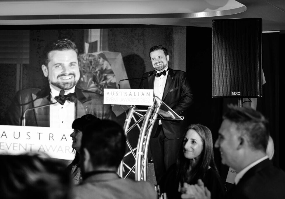 Nathan Cassar, MC smiling behind a lectern with the words "Australian Event Awards" written on the front, with his face mirrored behind him on a big LED screen.